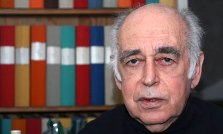 An old man with bushy black eyebrows in front of a bookshelf apparently holding arch binders.