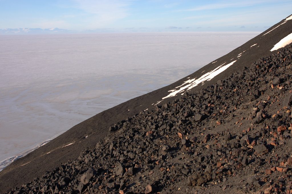 The long steep scree slope of Arrival Heights, plunging into McMurdo Sound at roughly a 45° angle. The Western Mountains are just visible as a faint blue on the horizon. The frozen sea is a slightly rosy grey, getting darker as it comes towards the land. The slope of the hillside is covered in medium-sized lava rocks, mostly black but with a few smaller reddish ones. A few patches of snow light up the further reaches of the hillside.