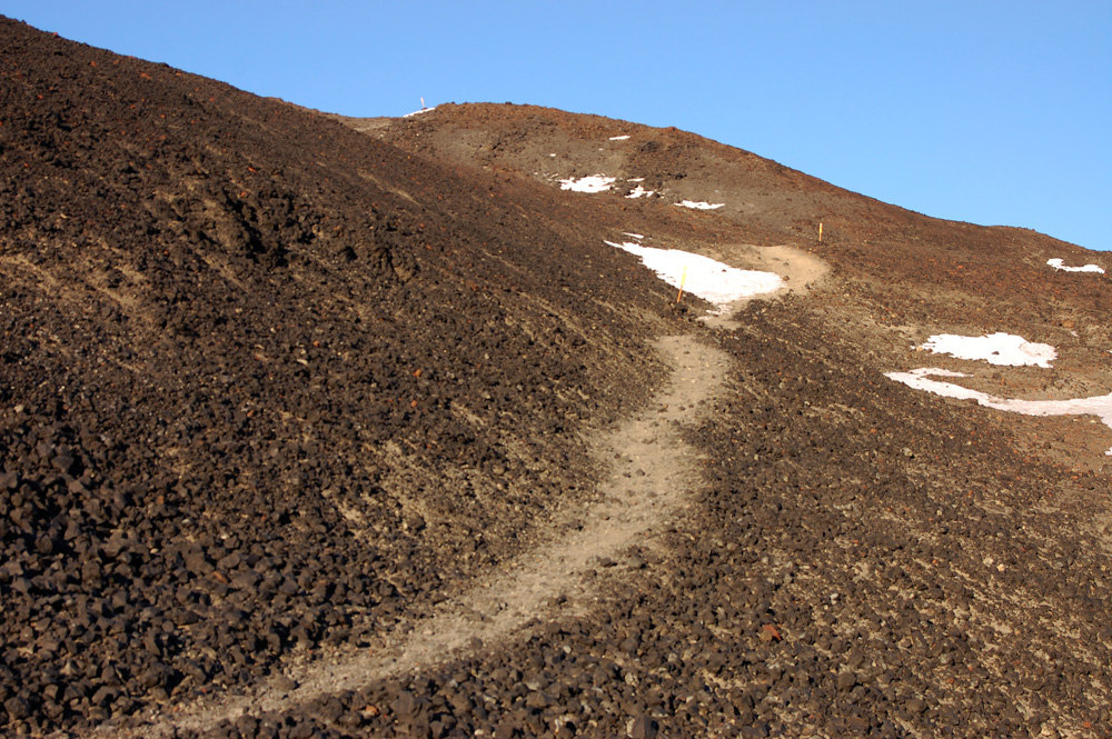A light dirt trail snakes up a dry rubbly slope towards a further hilltop with some rock outcroppings, all a sort of milk chocolate colour. Some small patches of snow huddle in hollows, and the sky is a radiant periwinkle.