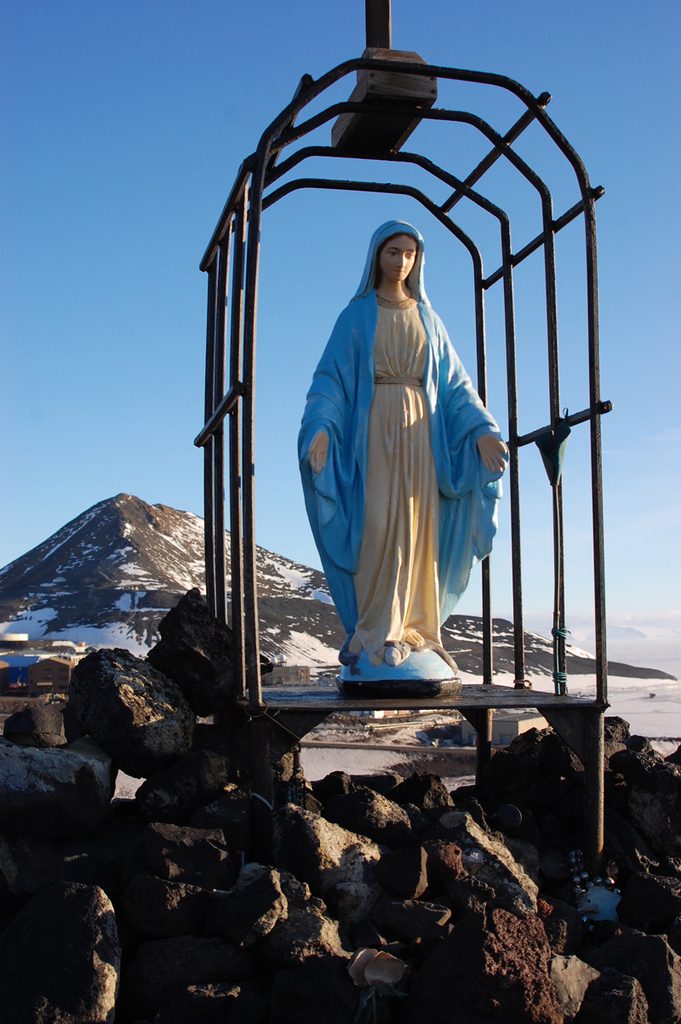 A traditional Catholic stautette of the Virgin Mary, in white and blue robes, stands in a grotto made of iron pipes, which sits on a small cairn of lava rocks on a low ridge of Arrival Heights. Observation Hill is visible in the background, under a stunning blue sky.