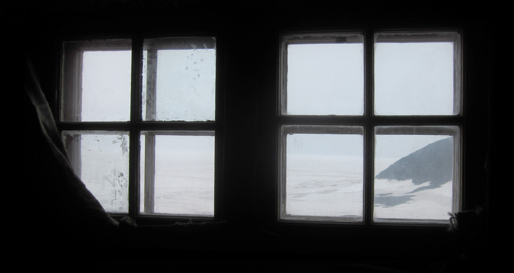A view out the windows of the pantry room of the Discovery Hut, towards the promontory which hosts Vince's Cross, though the cross is not visible.  The windows are very dusty, somewhat obscuring the grey day outside.  The interior of the hut is black, but a little bit of sacking catches the light to the left of the leftmost window.