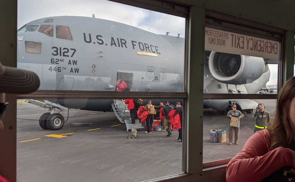 Passengers board an Antarctic-bound USAF C-17, receiving bagged lunches before proceeding up the gangway to the entrance hatch. Seen through the windows of a school bus.