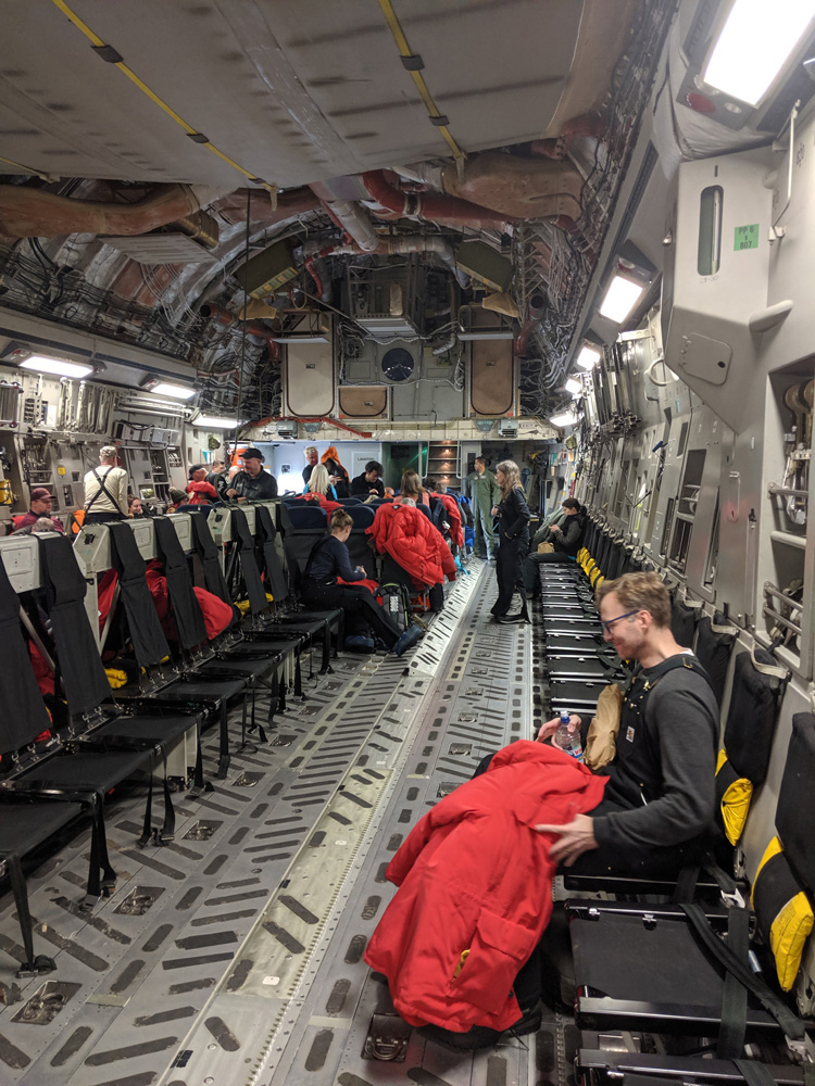 Looking forward in the passenger section of the hold of an Antarctic-bound USAF C-17. There is a utilitarian exposed metal floor, seats running the length of the cabin, and much exposed writing and ductwork on the walls and ceiling. One passenger sits in the foreground with his red parka on his lap.