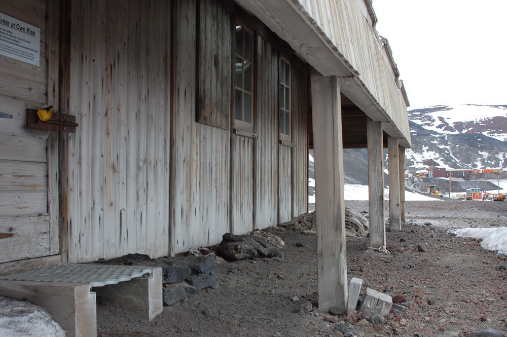 The side of the Discovery Hut facing Winter Quarters Bay.  Some of McMurdo's more northerly buildings are visible in the far background.  At the extreme left foreground is the entry door with its yellow lock and a sign declaring that one enters at one's own risk.  The building proceeds right from there with two windows, protected by the awning which is supported on its sturdy beams.