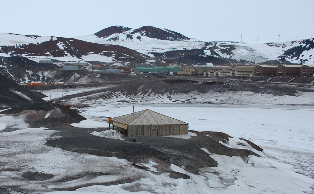 Looking over the Discovery Hut to McMurdo Station, showing frozen Winter Quarters Bay, the wharf, the road to Hut Point and the branch road to the wharf, most of the dorms, and Crater Hill rising behind it all.  The rock is covered with patches of dirty snow and the cloudy sky casts everything in a cold grey.  On the ridgeline to the right, three windmills harvest energy for the base to offset the diesel generator.