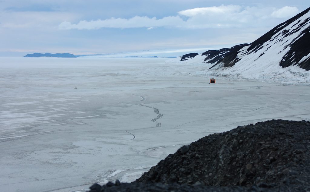 View of Cape Evans from Hut Point.  At the horizon, the Dllbridge Islands stand blue-grey against the white slope of Erebus down to the sea.  One scar of exposed rock at centre is The Ramp of Cape Evans.  Between there and here is the frozen sea with clearly visible cracks.  An orange fish hut sits near the base of Arrival Heights to the right.  The dark volcanic ridge of Hut Point is seen in the foreground, lower right.
