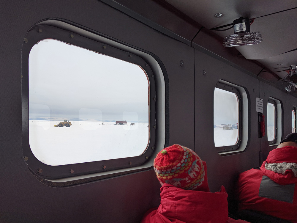 View out the windows of the big red people-mover, onto the heavy machinery at Phoenix Airfield, Antarctica, on a cloudy day.