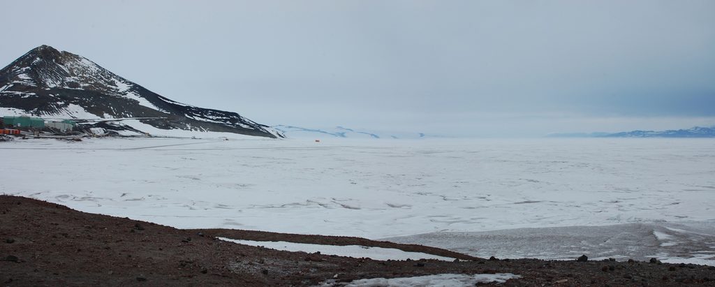 A panoramic shot of the southern view from the Discovery Hut.  Observation Hill rises on the left, with the lower buildings of McMurdo Station just visible below it.  Across the frozen sea ice one can make out White Island, its stripes of exposed rock the only thing delineating it from the grey sky, and further along, Minna Bluff and Black Island in gradating tiers of blue-grey.  The volcanic rubble of Hut Point is at your feet in the foreground, and the near ice is dirty with dust blown off the nearby hills.