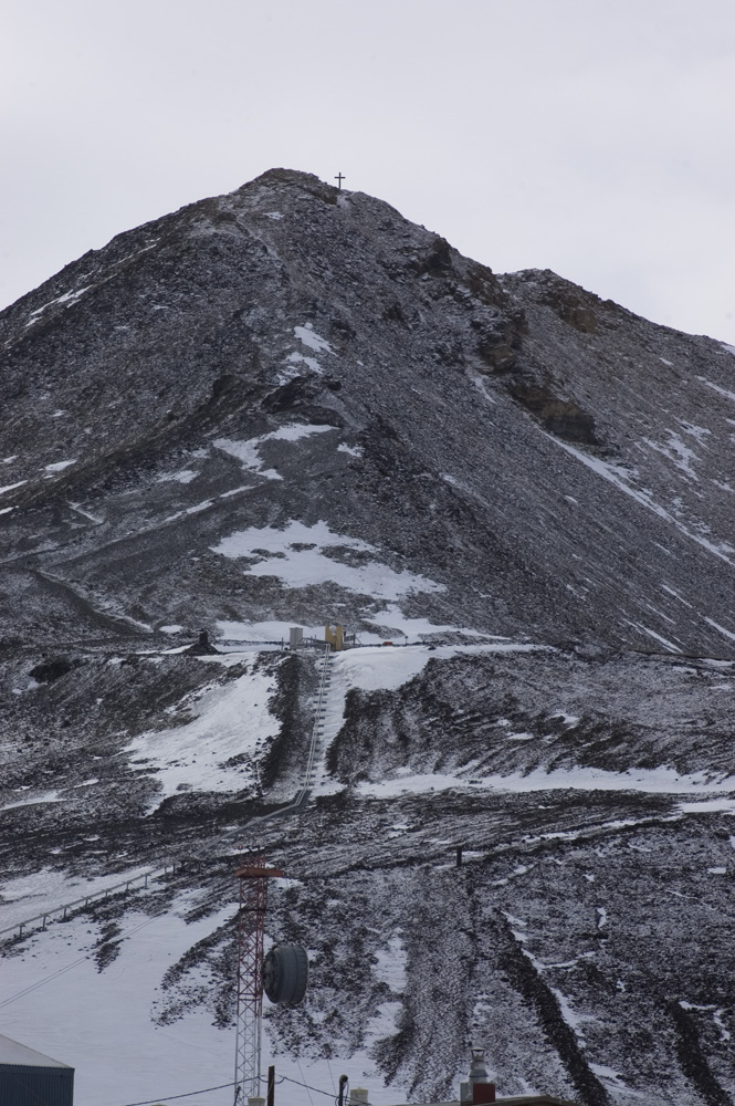A large cark conical hill topped with a tiny cross looms into a rose-grey sky. Patches of snow cling to its lower reaches, and the whole is lightly dusted with a fresh snowfall which makes the outcrops of bare rock on the right side stand out.