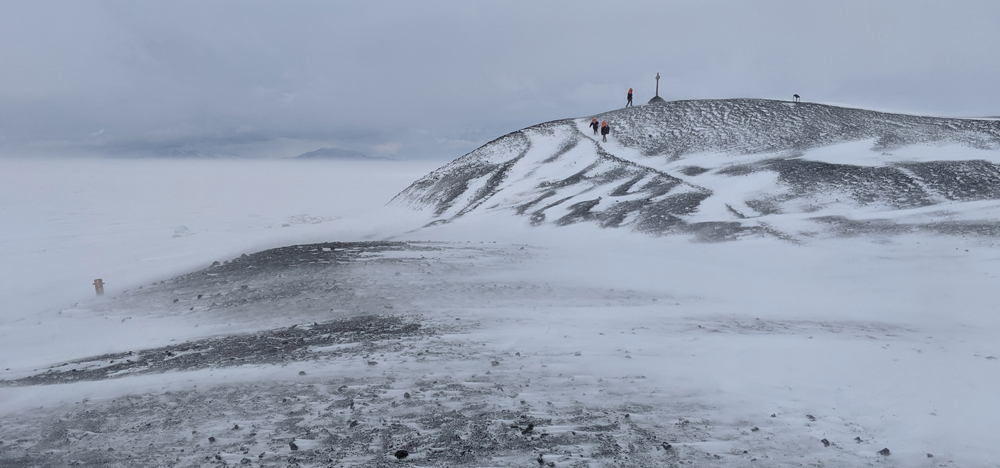 Some members of Antarctica New Zealand visit Vince's Cross on Hut Point, on a grey day with low drifting snow skidding across the dark gravel and leaving little drifts behind each exposed pebble.  In the distance, on the other side of the Sound, the clouds grow blue-grey towards the horizon, where the feet of the Western Mountains can be seen.