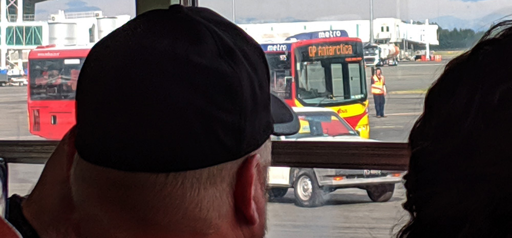 A Christchurch city bus with 'OP ANTARCTICA' on the LCD display indicating the route, seen over the shoulder of a man in a ball cap.