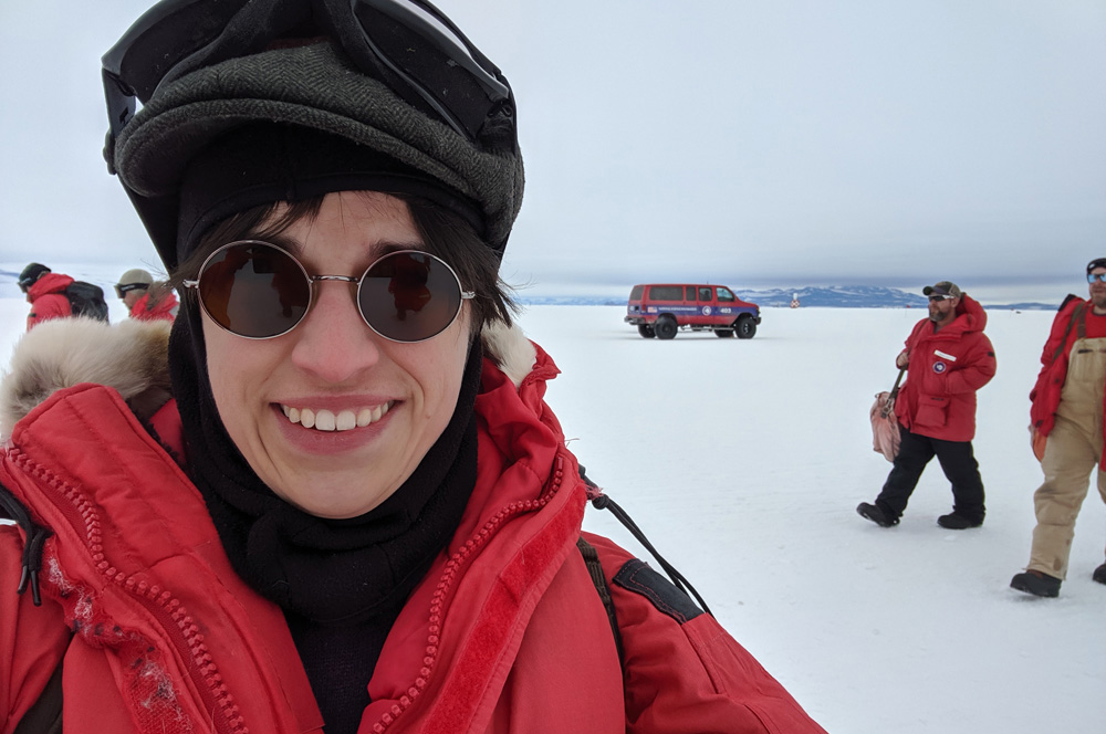 The author on the ice shelf at Phoenix Airfield, in sunglasses, green flat cap, and big red parka.  A few other passengers, an NSF shuttle, and Black Island in the background.