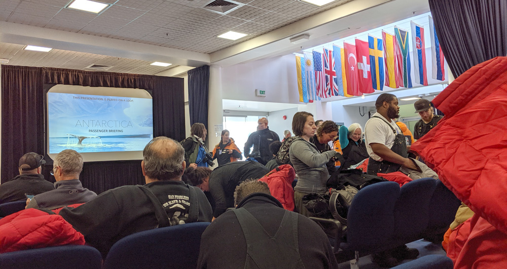A crowd of people find seats in a small conference room, to watch a video projected on a screen at the front, whose title card reads 'Antarctica'.  A number of flags hang above the hallway visible outside the room.