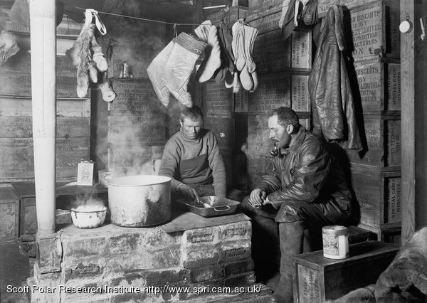 A monochrome photo of two men, one yonger and one middle-aged, surrounded by walls made of packing cases, as they cook on a knee-height brick stove. Various mitts and socks hang from a clothesline above.