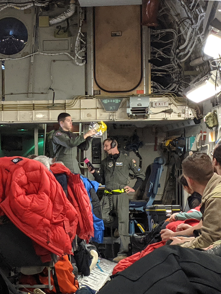 Two members of the NY Air National Guard do the safety briefing on an Antarctic-bound C-17. The gangly white one stands nonchalantly on the floor, reading out of a small booklet, while the shorter Latino one stands on a chair and demonstrates use of the oxygen system.