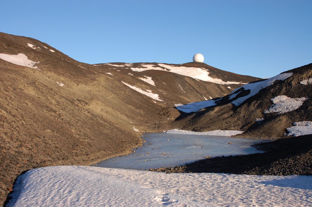 A view towards the primary white radome on Arrival Heights, through a gully in the brown hills with a frozen pond at the bottom. There is a patch of old dirty snow in the foreground with a few footprints in it. The sky is a piercing blue, which is reflected in the shadows on the snow.