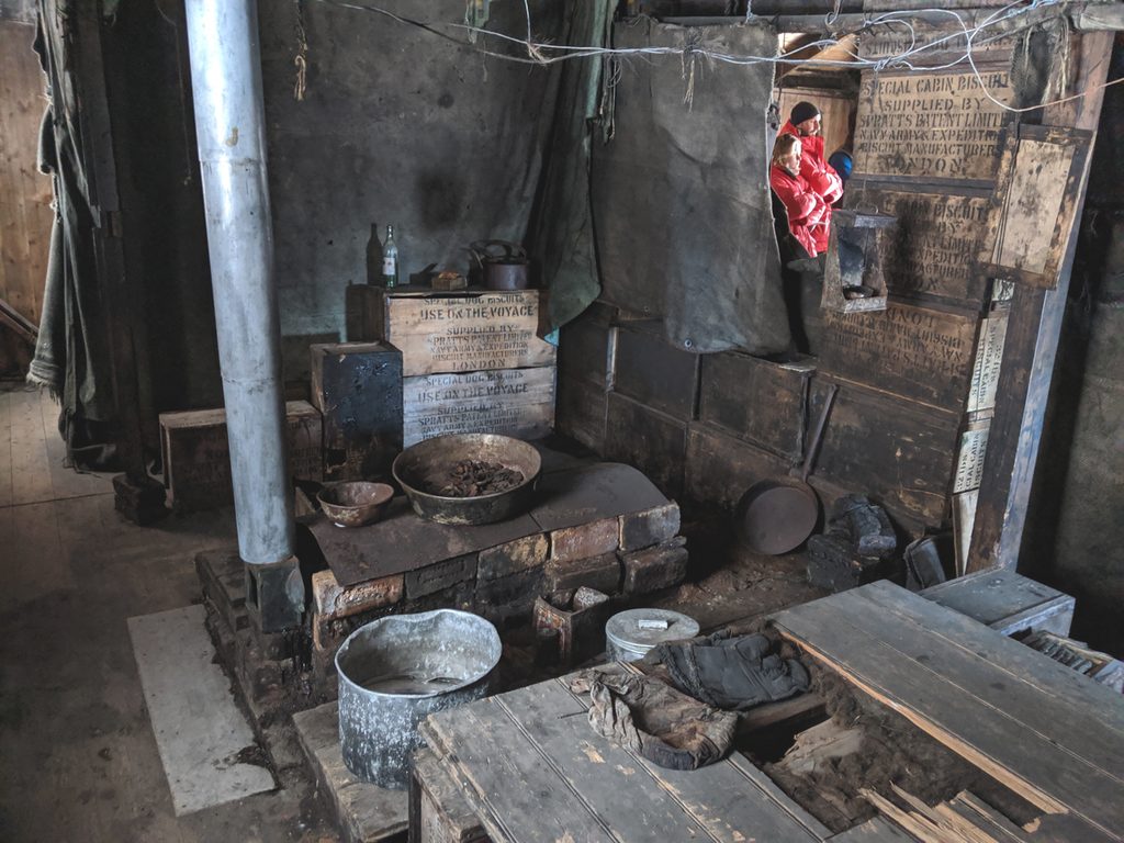 The cooking area of the Discovery Hut, view from the elevated sleeping platform towards the stove.  On and around the stove are a selection of pots and pans, and the zinc stovepipe climbs to the ceiling from the left side.  Some crates in the background serve as a counter.  On them is steniclled 'special dog biscuits: use on the voyage'.  The far wall of the kitchen is a sooty grey-green fabric; the wall to the right is made of packing cases.