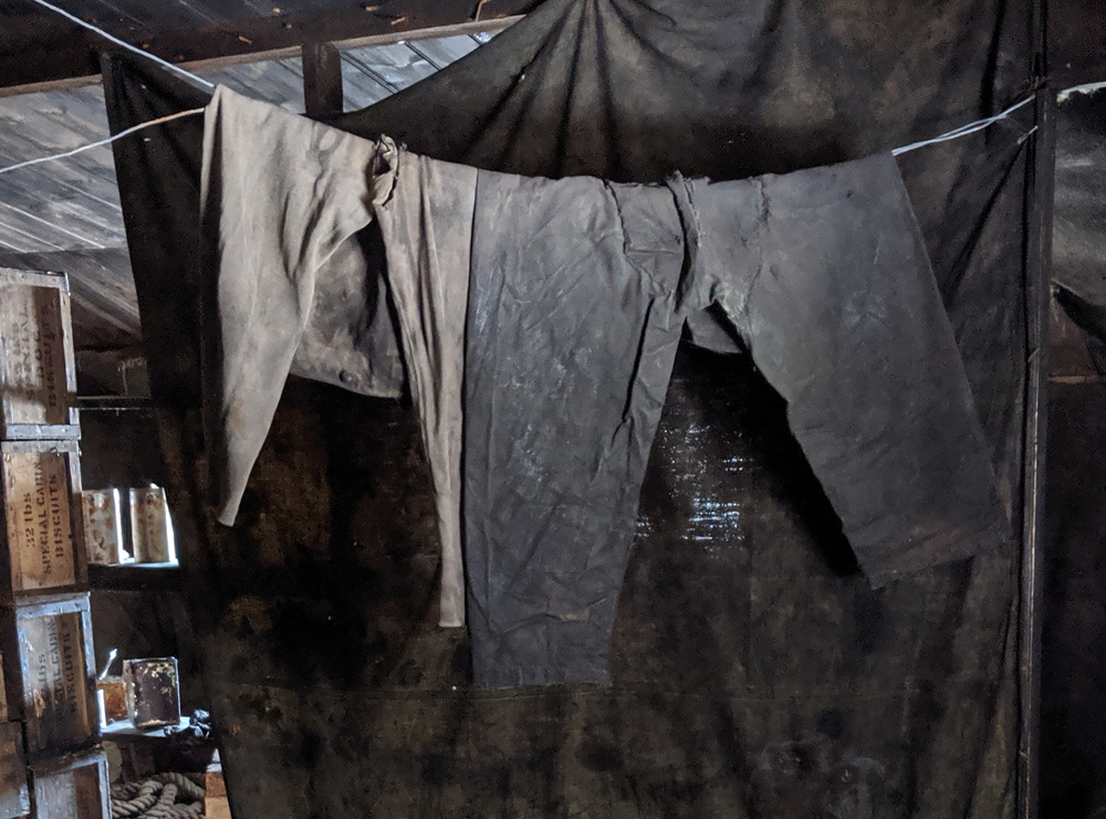 Two pairs of trousers hang on a line in the Discovery Hut kitchen area.  On the left is a pair of thick woollen long underwear, with a tear in the crotch and a waist button visible.  They may once have been beige but have been pretty heavily stained with soot into a warm grey.  To the right of these are a pair of very stiff-looking dark grey trousers which look homemade, with a large patch in the crotch area.  These may have been some of the winter wear made by the Ross Sea Party out of a discarded canvas tent.  They hang in front of a dark curtain and are lit by a window unseen to the right and behind the viewer.