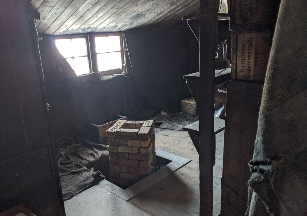 A view of the pantry area of the Discovery Hut from near the entry.  A hole has been built into the floor to accomodate a hollow square stack of bricks, offset diagonally to the square hole.  This served as an earth-anchored base for the pendulum experiments which measured gravity.  The ceiling slants down to the far wall which is hung with hessian, partly obscuring the window, and the larder shelves are visible to the right.