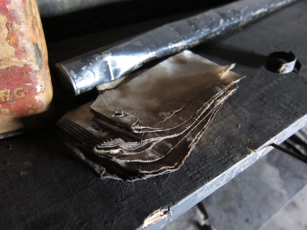 Closeup of a stack of tags on a shelf in the pantry of the Discovery Hut.  They are made of a stiff white fabric which has been saturated and stained brown by some sort of grease, perhaps.  Behind them is a bent aluminium tube and the corner of a baking powder tin.