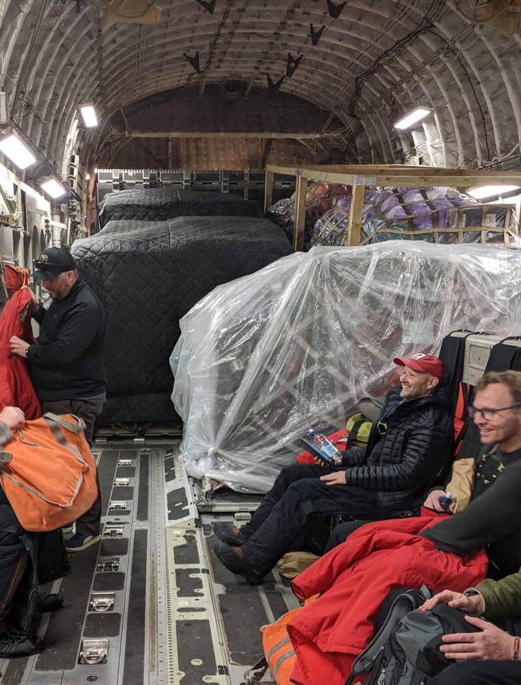 Looking aft in the hold of a C-17, very large blocks of cargo wrapped in plastic and cargo straps fill the rear of the fuselage. In the foreground some seated passengers are laughing and talking.