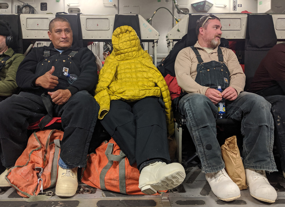 Three people sit in the centre seating on a C-17 bound for McMurdo. From left to right: a Native American man doing a thumbs-up, a woman leaning back in her seat with a yellow puffy jacket covering her head and torso, and a middle-aged white man chatting to someone unseen off to his side.