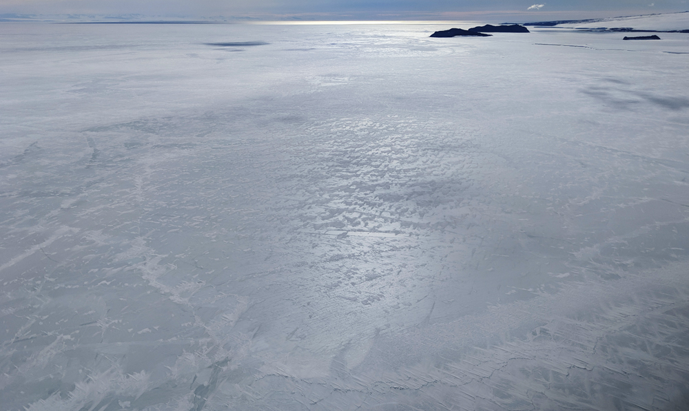 A helicopter shot of the sea ice south of Cape Evans.  The horizon is very high in the frame, affording a full vantage of the texture of the rozen surface of McMurdo Sound, mostly a medium greyish green but criss-crossed with white snowdrifts.  The exposed ice shines under the sun which is directly in front of us, giving the whole flat surface the feel of a billiard ball.  At the very farthest strip of the horizon, open water reflects a strip of bronze to the sky.