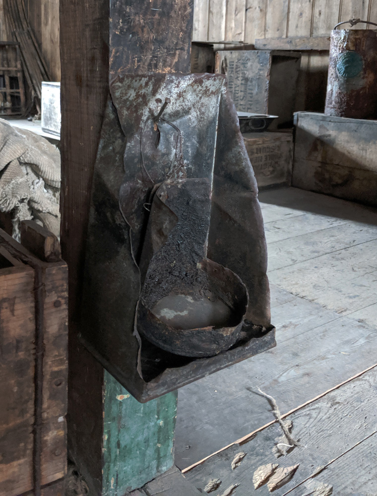 An improvised metal cnotraption is nailed near the bottom of one of the roof support beams in the Discovery Hut.  It looks like a rectangular box has been cut diagonally, the long flat side against the beam and the angled sides sloping out from it.  On the bottom of the box is an open cylinder with some waxy residue on the bottom.  The whole thing is very sooty and greasy looking.