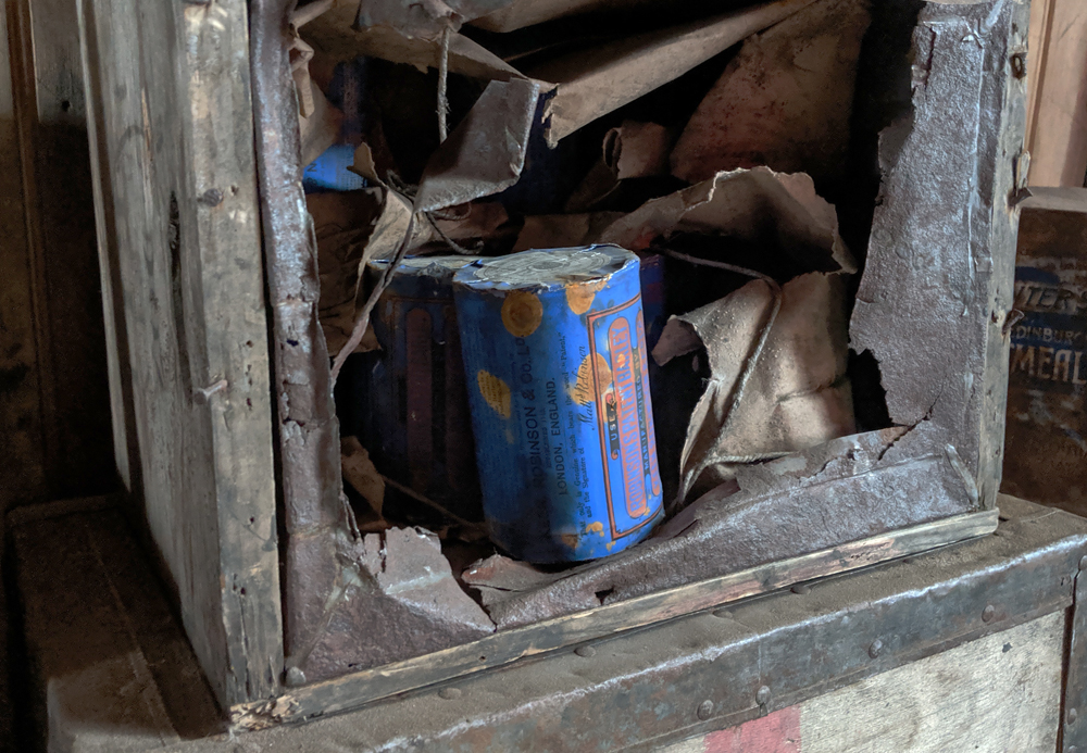 Inside a wooden crate on its side are some tins with an ultramarine label, which reads 'Robinson's Patent Barley' in red.  There are orange splotches of rust on the label.  The crate had once had a thin metal sheet covering it but this has been torn away to reveal the tins.