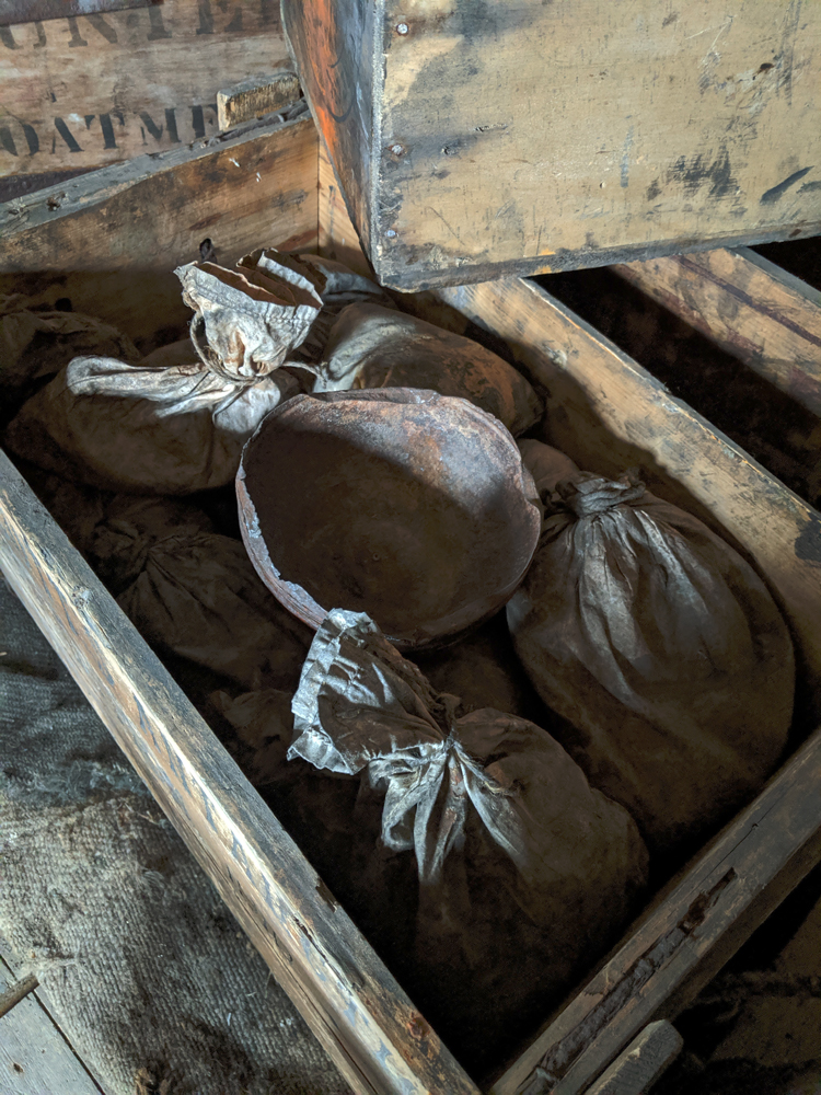 Inside an old crate at Discovery Hut, some grubby muslin bags tied with string at the neck, with a hemispherical metal bowl resting on top of them.  A portion of a crate in the background reads 'oatmeal' in stencilled letters.