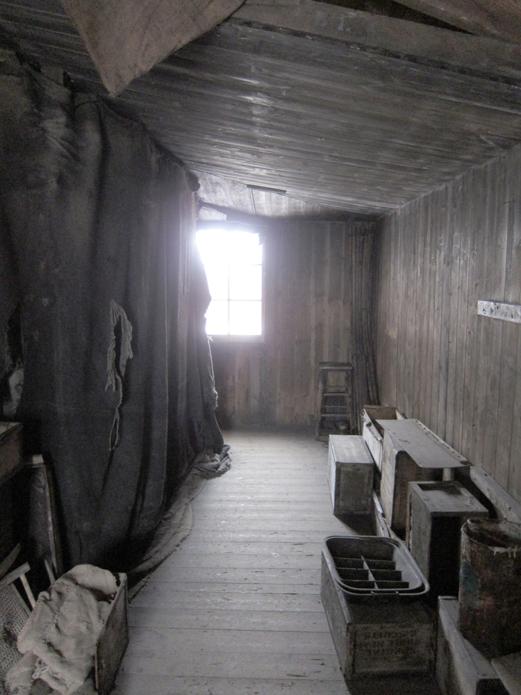 Looking down the passageway from the main space of the hut around to the kitchen area.  There is a very bright window at the end of it, which sheds its light on the rough curtain, forming the left wall of the passage, and an assortment of crates pushed against the exterior wall of the hut, forming the right.