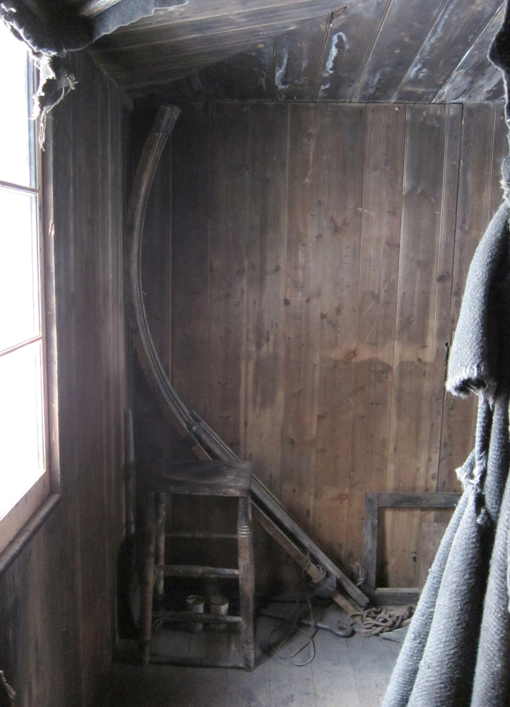 A set of curved wooden staves is propped up against the corner of the Discovery Hut, behind a square wooden stool.  They are attached at the top, and if fanned out would make a shape like one of those old-fashioned Christmas bell decorations made of cardboard fins with webbed tissue paper in between, minus the bottom flange.