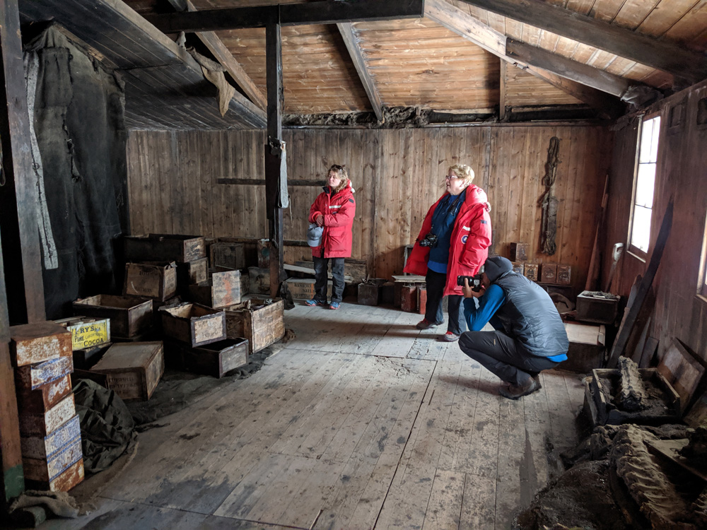 The main room of the Discovery Hut, as seen from the entrance.  Light from two windows on the right-hand wall illuminates a space about 15 feet wide and 30 feet long, with walls about seven feet high, a wooden roof with exposed beams sloping gently upwards from there to a central peak out of frame.  On the right, under the windows, are some old butchered seal carcasses, a large wooden box, and some other wooden bits.  On the left are some loosely stacked biscuit cases and, next to the nearer roof support beam, a neat stack of Huntley & Palmer biscuit tins.  Behind these is a curtain of rough material, stained black my blubber soot.  This open space wraps around the corner of the division made by the cases and curtain, out of sight to the left.