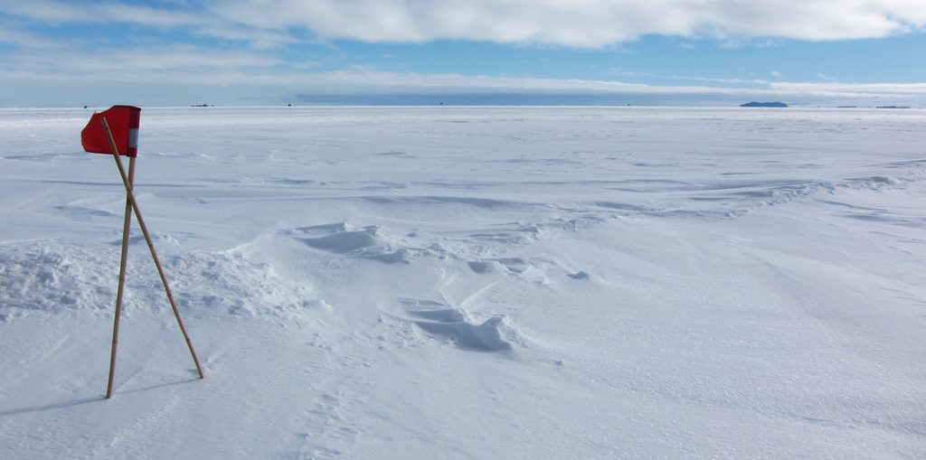 Looking north up McMurdo Sound, covered in ice and drifted snow.  A distant cloud bank shows a dark patch underneath, where it is reflecting the open water off Cape Royds.  Tent and Inaccessible Islands can be seen on the horizon at upper right.  In the foreground, two crossed bamboos with red flags indicate a crossing of a known crack.